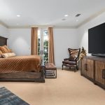 A spacious bedroom with a large bed, brown bedding, wooden nightstands, a dark armchair, a long wooden dresser, and a large flat-screen TV. Light walls, beige carpet, and sunlight streams through tall windows with peach curtains.