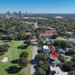 Aerial view of a suburban neighborhood with a red location marker over one house, a golf course on the left, and a city skyline in the background under a clear blue sky.