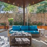 Covered patio with a wicker sofa featuring blue cushions, two matching armchairs, and a wicker coffee table, set on a tiled floor. The area is surrounded by planter boxes, greenery, and a brick privacy wall.
