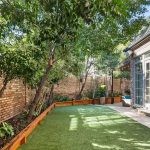 A private backyard featuring green artificial grass, raised wooden garden beds with plants, brick walls, and a house with large French doors and tall windows.