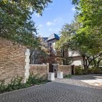 Brick house with a patterned circular driveway, tall trees providing shade, and a small gate leading to the front entrance. Blue sky is visible above the roof and leafy branches frame the scene.