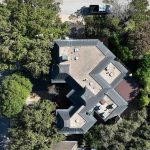 Aerial view of a large, uniquely shaped building with a flat roof surrounded by trees and greenery, located at the corner of two streets in a residential neighborhood. Cars are parked along the road.