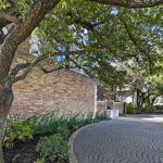 A curved stone driveway lined with green plants and shaded by tall trees runs alongside a brick building on a sunny day.