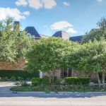 A brick building is partially hidden behind several leafy trees and shrubs, with a well-maintained garden bed in front. The sky is bright and partly cloudy.