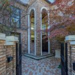 A brick courtyard with open iron gates leads to a grand entrance featuring tall arched doorways, large windows, and a tree with red leaves on the right side.