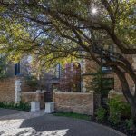 A large brick house is partly shaded by tall trees with spreading branches. Sunlight filters through the leaves, casting dappled shadows on the driveway and the garden area beside the house.