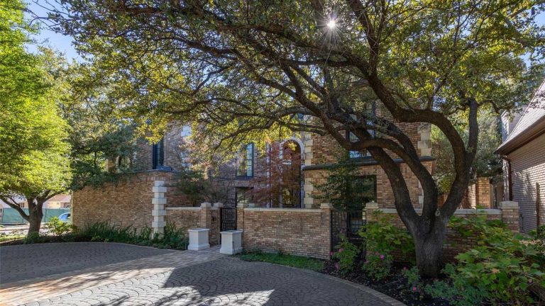 A large brick house is partly shaded by tall trees with spreading branches. Sunlight filters through the leaves, casting dappled shadows on the driveway and the garden area beside the house.