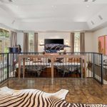A spacious, elegant living room features animal-themed decor, including a zebra-striped rug, animal sculptures, and a mounted animal head. Large windows and light curtains provide natural light, while parquet floors and art adorn the walls.