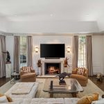 A modern living room with large windows, beige sofas, a fireplace under a wall-mounted TV, two armchairs, decorative sculptures, and two large pink cactus-themed artworks on the wall.
