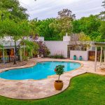Modern house with large windows and a kidney-shaped pool surrounded by stone tiles. There’s a small pergola with lounge chairs and a grassy yard bordered by dense green trees. A potted plant sits near the pool’s edge.