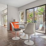 A modern dining area with white chairs and a round table next to large glass doors, offering a view of a backyard pool and trees. The space features gray tile flooring and an orange cabinet.