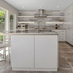 Modern kitchen with white cabinets, stainless steel appliances, a large island with a double sink, two metal chairs, and large windows letting in natural light. The backsplash features gray and white horizontal tiles.