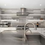 Modern kitchen with stainless steel island and sink, gray cabinets, a gas stove, range hood, floating shelves with dishes, and large windows letting in natural light. Striped gray backsplash adds a sleek, contemporary feel.