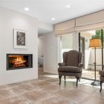 A modern living space with two patterned armchairs by a floor lamp, a fireplace with a framed picture above, large floor tiles, and a bedroom visible through an open doorway. Sunlight streams through glass doors to a patio.
