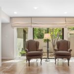 A cozy living room with two patterned armchairs by a window, a lamp between them, a fireplace on the left, tiled floor, and a view of a small garden outside.