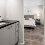 A modern bathroom with a black countertop and white cabinets opens to a bedroom with a gray upholstered bed, white bedding, a nightstand, lamp, and framed art on the wall. Natural light enters through a window.