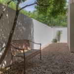 A narrow outdoor patio with gravel ground, a metal bench, white walls, potted plants at the far end, trees casting shadows, and large sliding glass doors on the right.
