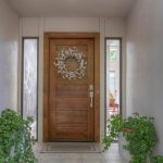 A wooden front door with a white floral wreath, flanked by two tall narrow windows, two potted green plants on either side, and a glimpse of a living room with an orange sofa inside.