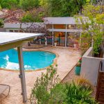 A modern backyard features a kidney-shaped swimming pool surrounded by stone tiles, lounge chairs, potted plants, and trees. The pool is adjacent to a contemporary house with large windows.
