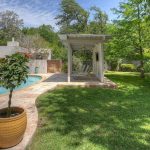 A backyard with a swimming pool, a potted plant on a stone patio, a grassy lawn, and a covered seating area with chairs under a white pergola, surrounded by lush green trees and plants.