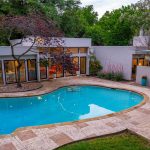 Modern house with large windows and a curved swimming pool in a landscaped backyard surrounded by trees and plants, with lounge chairs and string lights reflecting in the water.