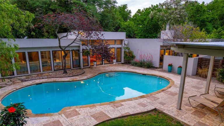 Modern house with large windows and a curved swimming pool in a landscaped backyard surrounded by trees and plants, with lounge chairs and string lights reflecting in the water.