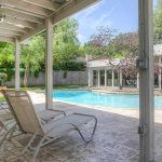 A covered patio with lounge chairs overlooks a backyard swimming pool, surrounded by trees, a fence, and a modern house with large windows. The scene is bright and inviting on a sunny day.