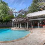 A backyard with a curvy swimming pool surrounded by stamped concrete, lush green trees, a flowering plant in a turquoise pot, and a modern house with large glass windows and doors.