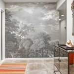 A hallway with a wooden door, gray tile floor, and a black-and-white wall mural of trees. A mirrored console table with decor and a striped rug are also present, along with a decorative mirror on the wall.