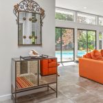 A modern living room with an orange sofa, large windows overlooking a backyard pool, a mirrored console table with a decorative mirror above it, and a hallway leading to other rooms.
