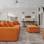 Modern living room with orange sofas and ottoman, gray tile floors, a fireplace, large flat-screen TV, wall art, decorative lamps on a console table, and glass doors opening to a backyard with a pool.