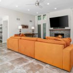 A modern living room with orange sofas, a fireplace under a wall-mounted TV, large windows with white curtains, and gray tile flooring. An open doorway leads to a kitchen with stainless steel appliances.