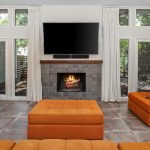 A modern living room with orange sofas, a matching ottoman, a tiled fireplace with a fire burning, a wall-mounted TV above it, large windows with white curtains, and a wooden desk with a lamp on the left.