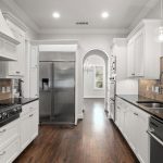 Modern kitchen with white cabinets, stainless steel appliances, dark countertops, a built-in gas stove, tile backsplash, hardwood floors, and an arched doorway leading to a dining room.