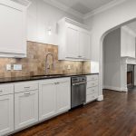 Modern kitchen with white cabinets, stainless steel appliances, a tile backsplash, dark countertops, and wood flooring. An arched doorway leads to a living area with a fireplace and French doors.