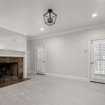 A bright, unfurnished room with light gray walls and carpet, a dark marble fireplace, recessed lights, a black geometric ceiling light, and two doors with white plantation shutters.