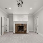 A bright, empty room with light gray walls, white trim, and carpet. A marble fireplace is centered on one wall, flanked by a door on the left and a glass-paned door with shutters on the right. A black ceiling light hangs above.