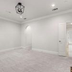 A bright, empty room with light carpet, white walls, a ceiling light fixture, and an open door leading to a kitchen with white cabinets and tile flooring. An arched doorway and fireplace are also visible.