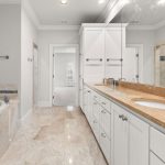 Spacious bathroom with a large soaking tub, double sinks on a long marble countertop, white cabinets, and floor-to-ceiling mirrors. The floor and tub surround are tiled in light beige marble.