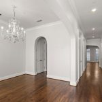 A spacious hallway with polished wooden floors, white walls, a decorative chandelier, arched doorway, and a wooden staircase with black railing leading upstairs. Natural light filters in from windows at the far end.