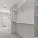 Spacious walk-in closet with white shelves, drawers, and hanging rods on light carpeted flooring; a door with shuttered blinds lets in natural light at the far end.