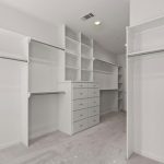 Spacious walk-in closet with white shelves, multiple hanging rods, and built-in drawers, all set against light-colored walls and carpeted flooring.
