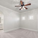 A bright, empty room with light-colored walls and carpet, a ceiling fan with lights, two small shuttered windows, a glass door with shutters, and an arched doorway leading to a bathroom with a double vanity.
