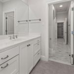A modern bathroom with white cabinets, a wide countertop with a sink and faucet, a large wall mirror, and a view into a walk-in closet through an open doorway. The space is bright with clean, neutral decor.