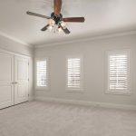 A bright, empty bedroom with light beige walls, two windows with white shutters, a ceiling fan with lights, double closet doors, and carpeted flooring.