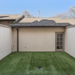 A small, enclosed outdoor courtyard with artificial grass, surrounded by high beige walls. There is a single door with glass panes leading into the building and a string of lights on one side.