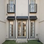 A beige townhouse exterior with three glass doors, two small balconies on the upper floor, black awnings, and artificial grass in a narrow fenced courtyard.