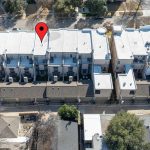 Aerial view of a modern apartment complex with white roofs. A red location pin marks one unit near the center of the top row, and surrounding streets and trees are visible.