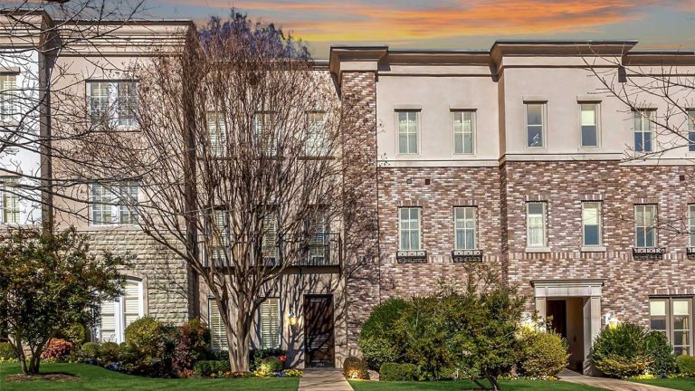 A row of modern townhouses with brick and stone facades, large windows, and trimmed landscaping, set against a colorful sunset sky. A leafless tree stands in front of one townhouse.