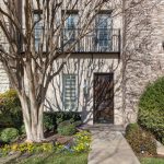 A townhouse entrance with a dark wooden door, large front window with shutters, leafless tree, trimmed bushes, and colorful flowers along a paved walkway against a stone and brick façade.